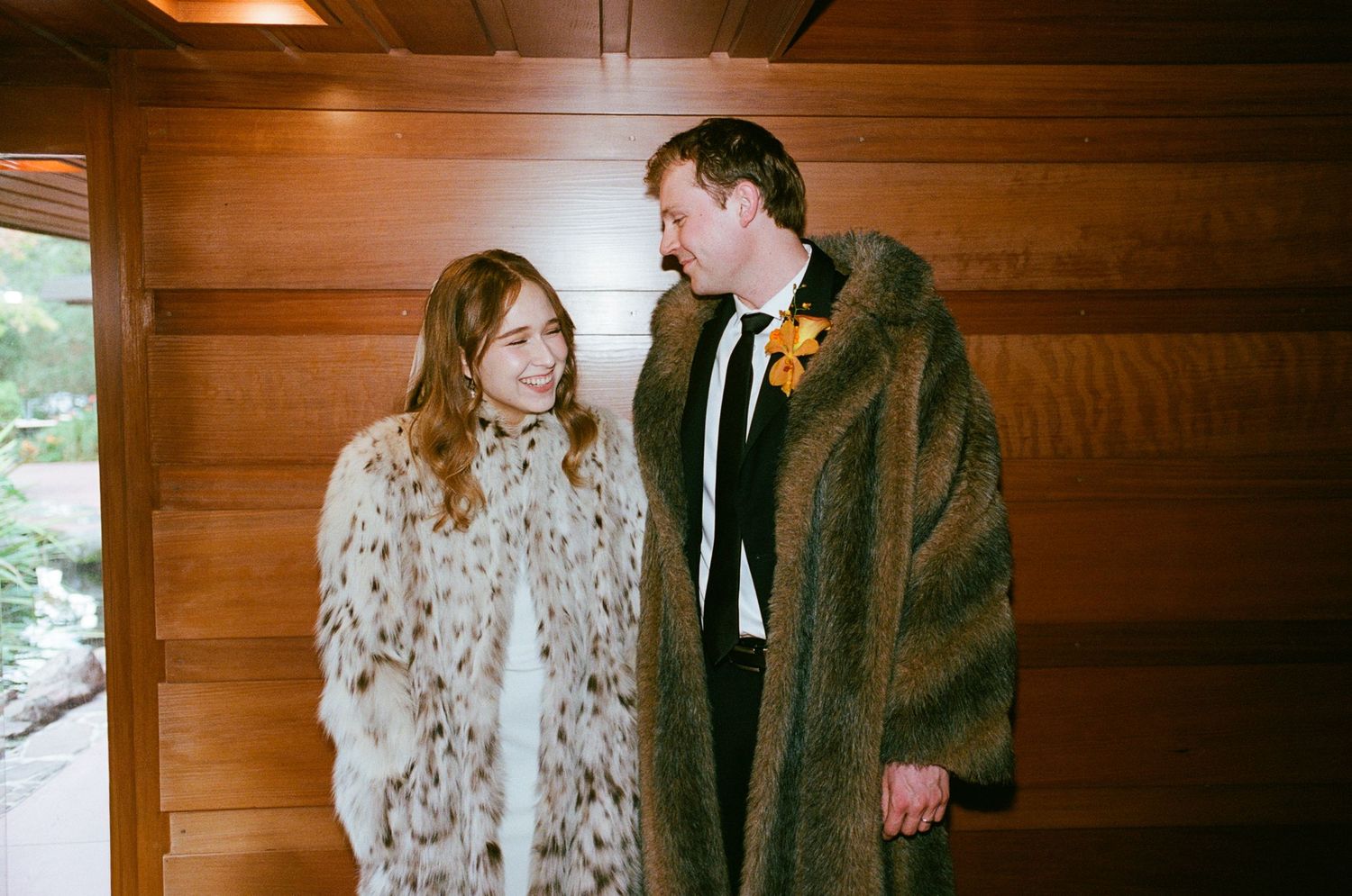 Bride and groom smiling at each other wearing matching oversized fur coats with a yellow boutonniere at a wood-paneled venue.