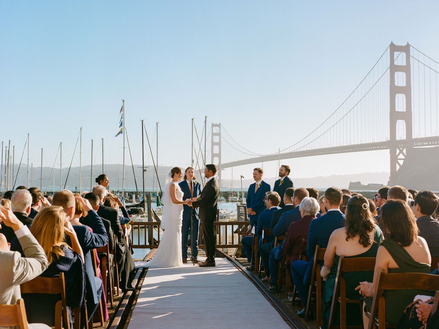 An outdoor wedding ceremony takes place on a boat deck with the iconic Golden Gate Bridge visible in the background.