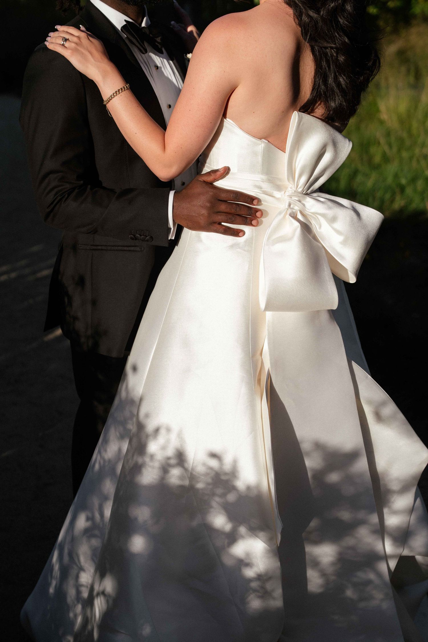 Bride in strapless white satin gown with large bow embracing groom in black tuxedo at outdoor wedding.
