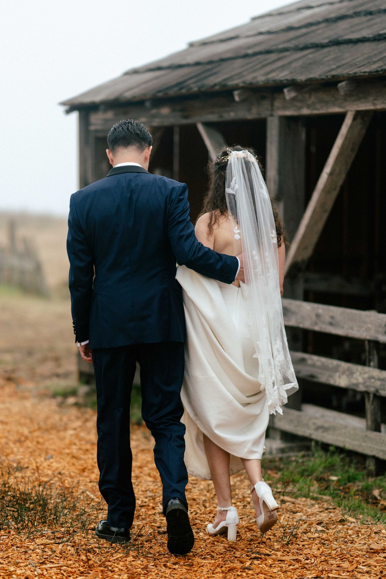 Bride and groom walking hand in hand toward a rustic wooden barn on their wedding day.