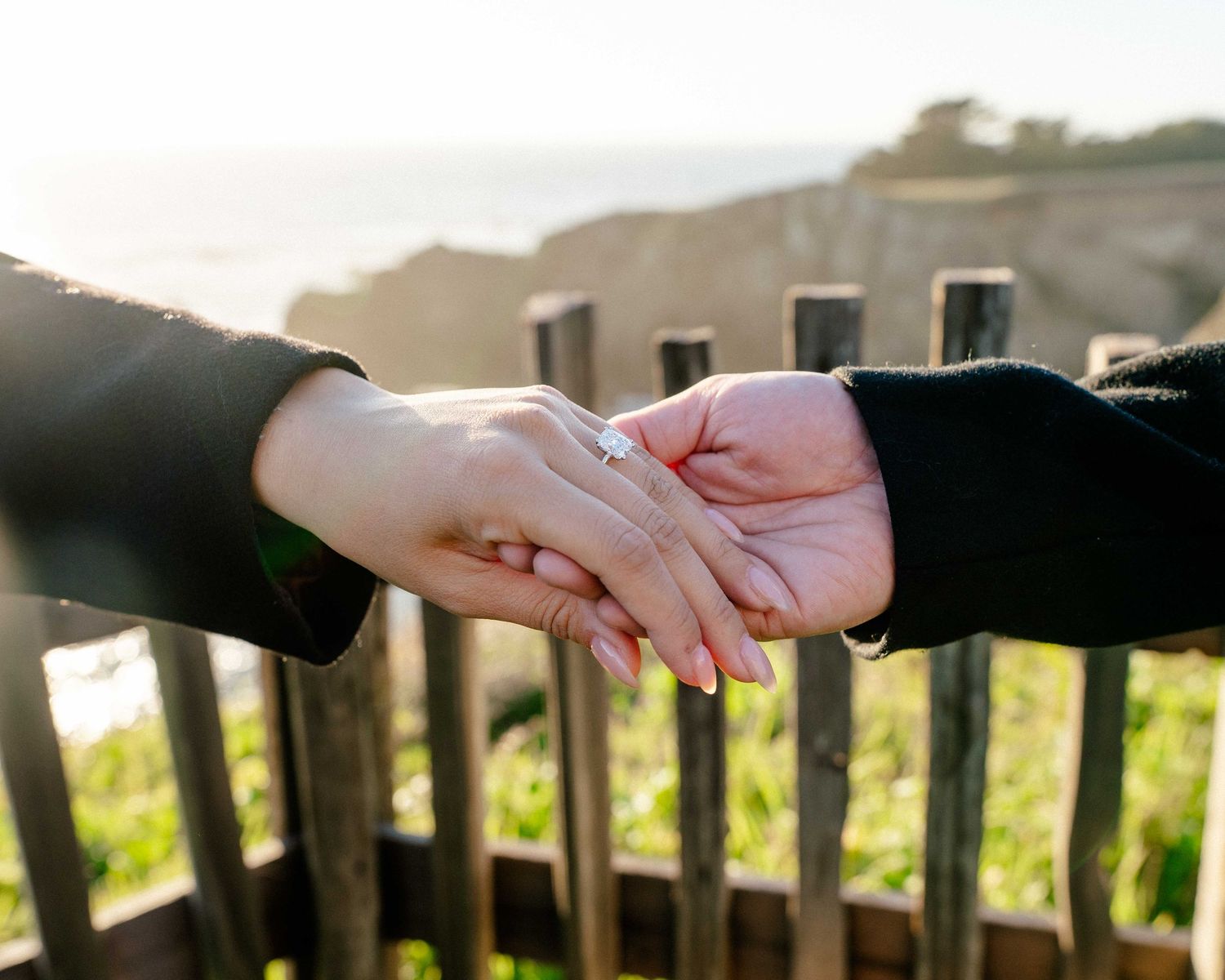Two hands holding near a wooden fence, one wearing an engagement ring, with a scenic coastal backdrop.