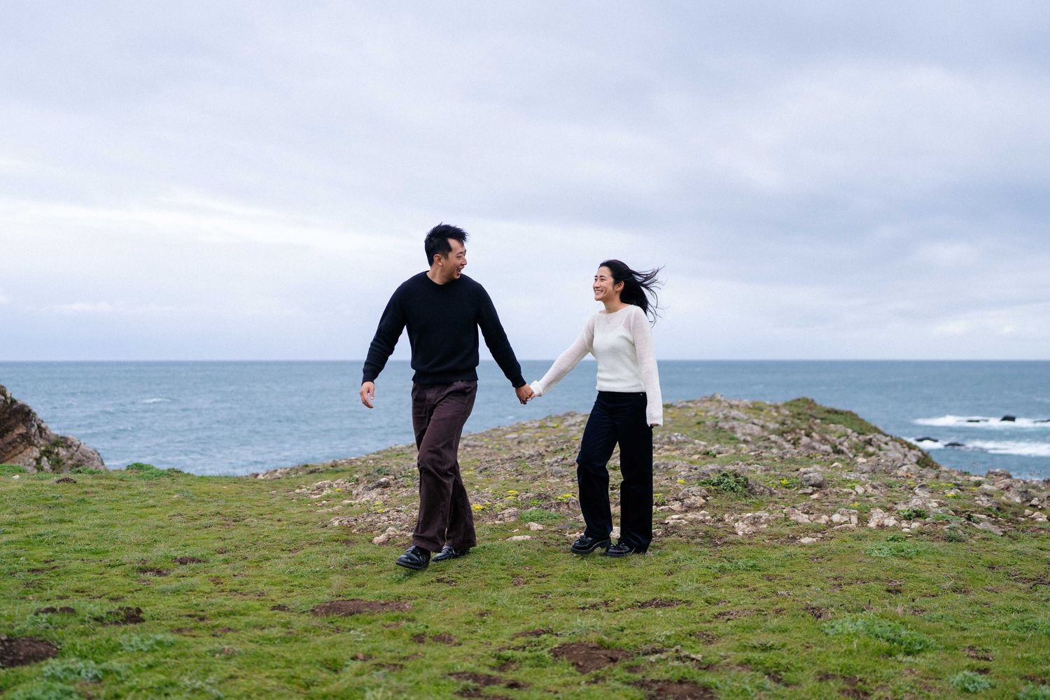 Couple holding hands on a grassy coastal cliff with ocean views under a cloudy sky.