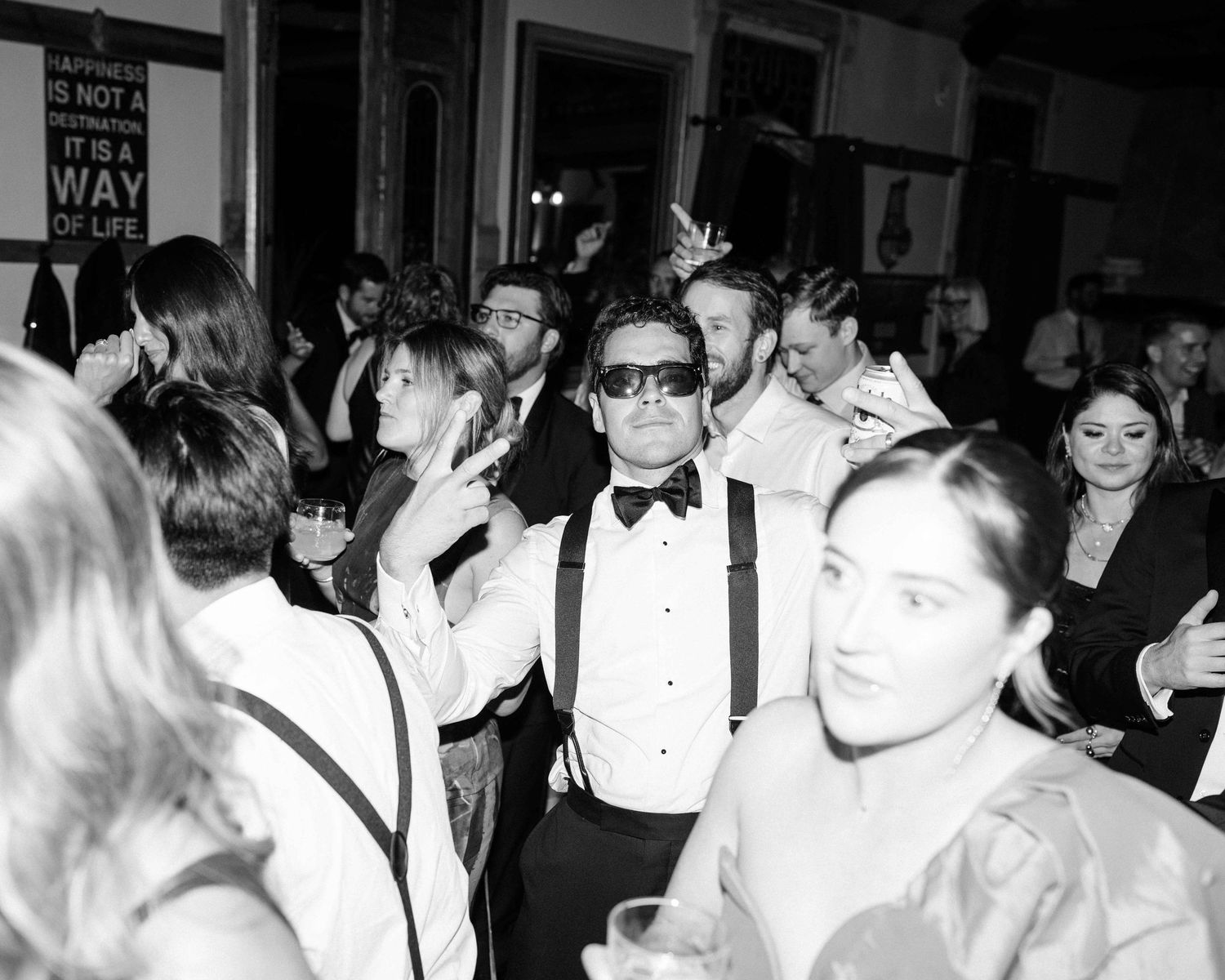 Elegant black and white wedding reception dance floor scene with guests celebrating, man in bow tie and suspenders at center.