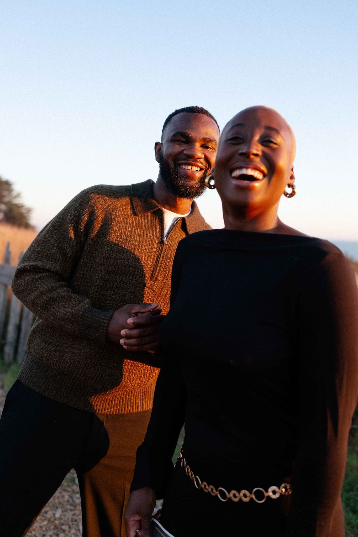 A laughing Black couple embraces outdoors at golden hour, radiating joy and warmth together.