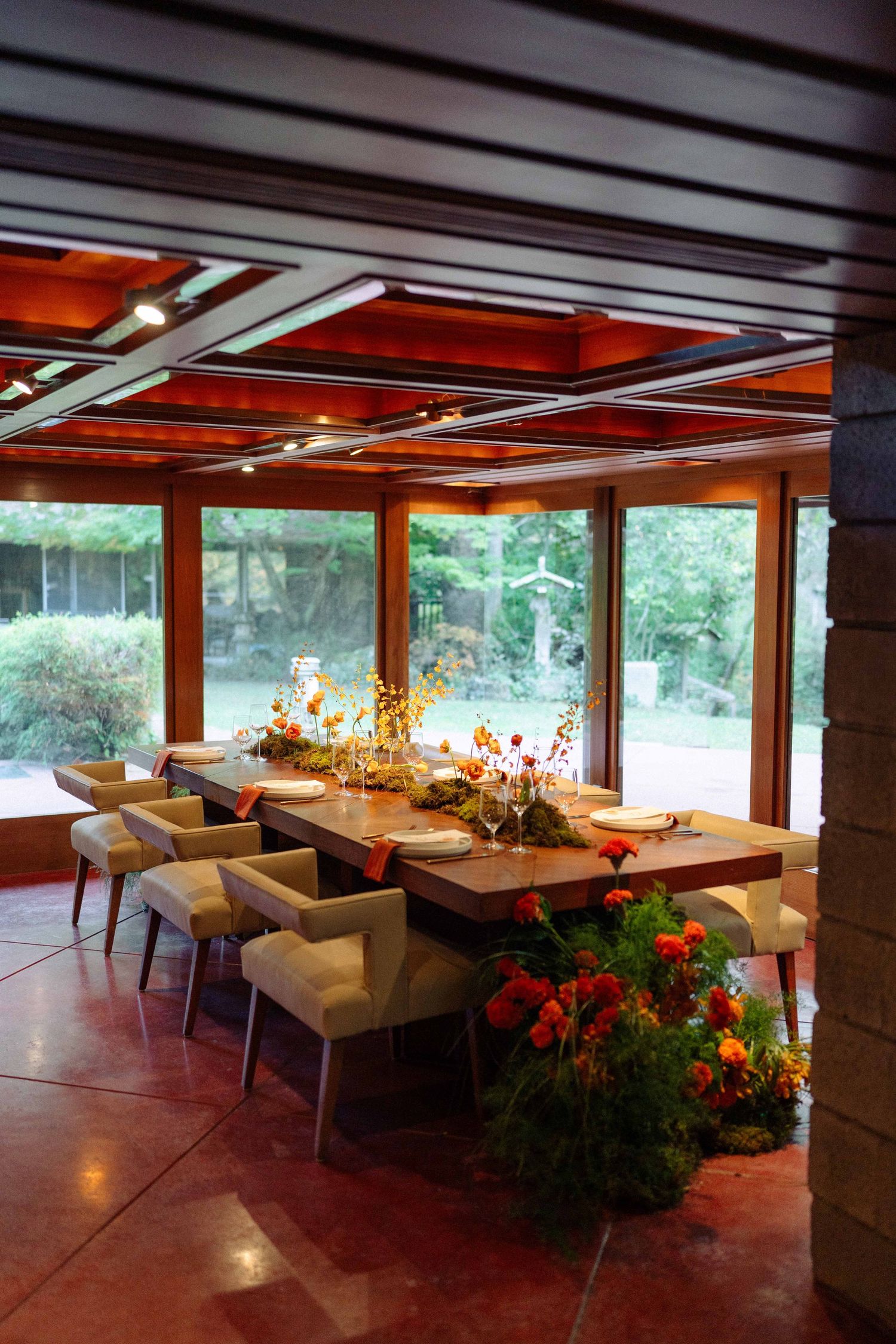 Elegant wedding reception dining table with orange floral arrangements in a mid-century modern glass-walled room.