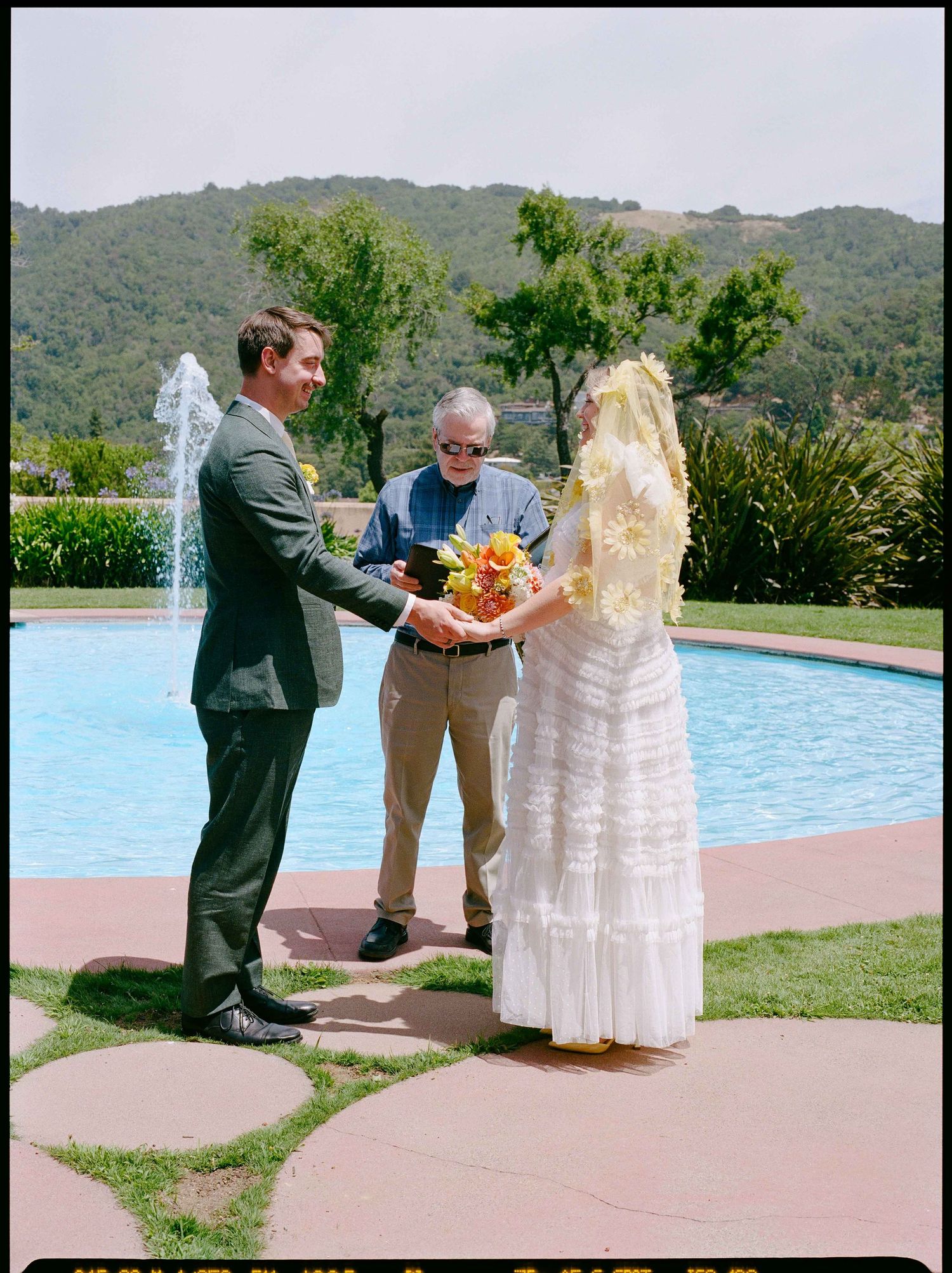 Outdoor wedding ceremony by a pool with a bride in white dress, groom in suit, and officiant amid lush green hills.