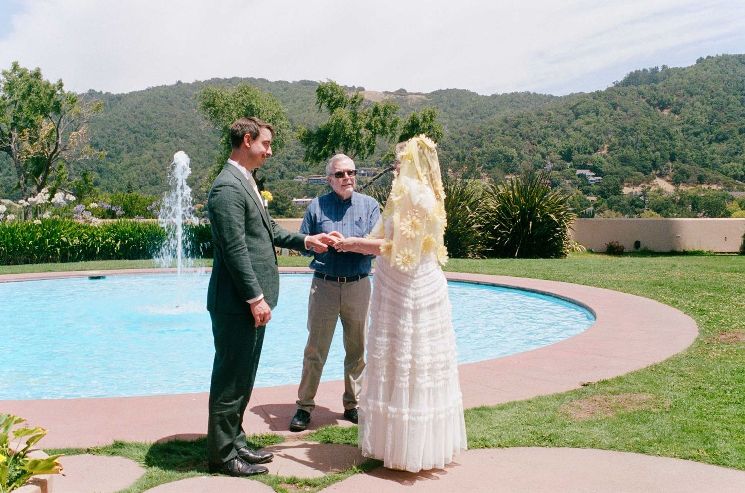 Outdoor wedding ceremony by a pool with green hills, couple exchanging vows with officiant, sunny day.