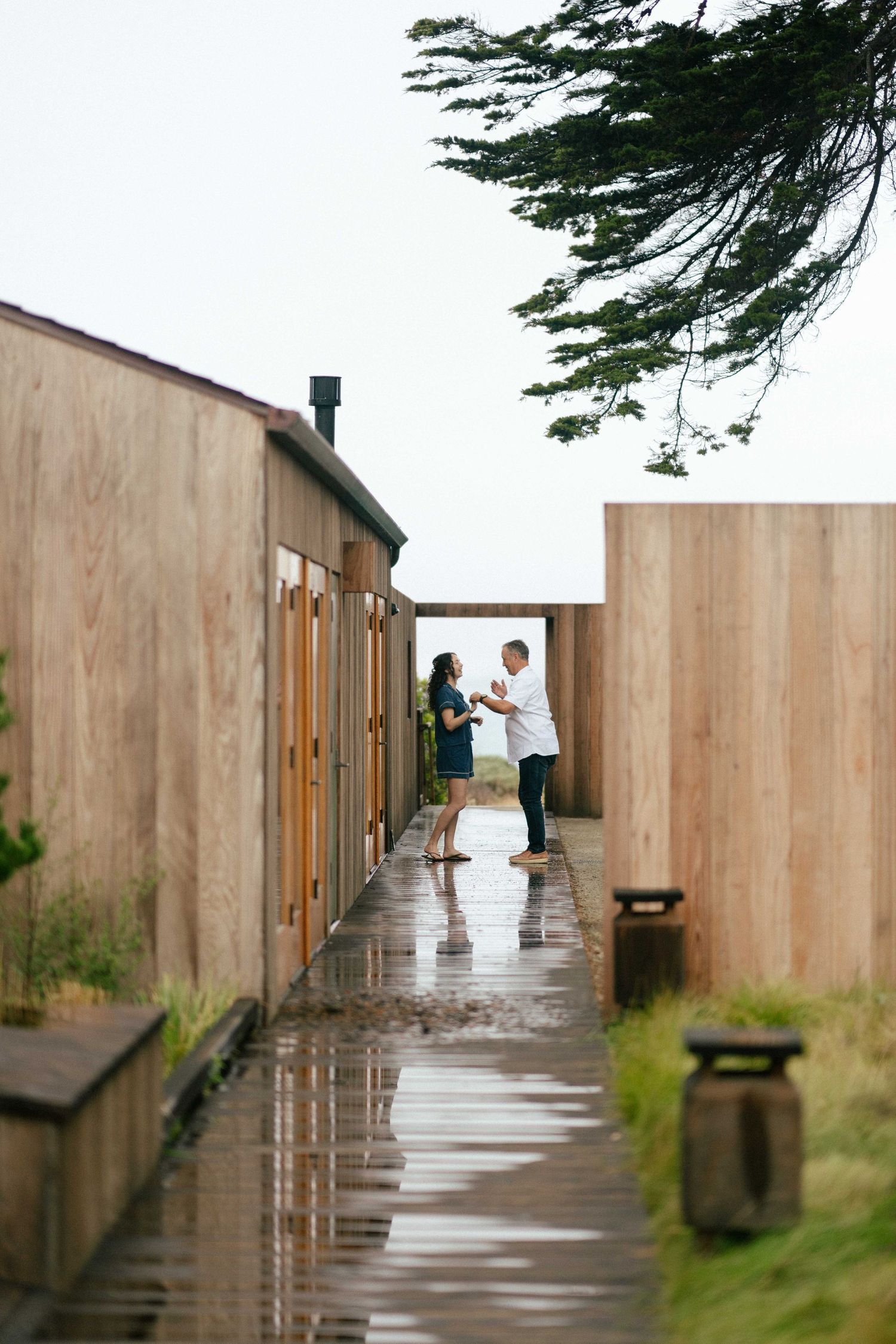 Couple sharing a romantic moment on a rain-soaked wooden walkway between modern timber buildings on a cloudy day.