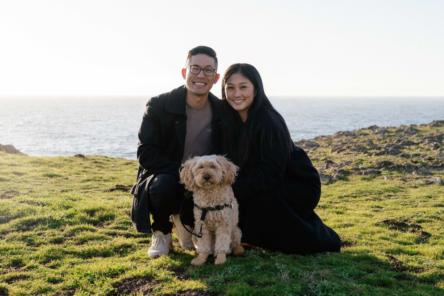 Couple smiling with fluffy dog on green coastal cliff overlooking ocean at sunset.