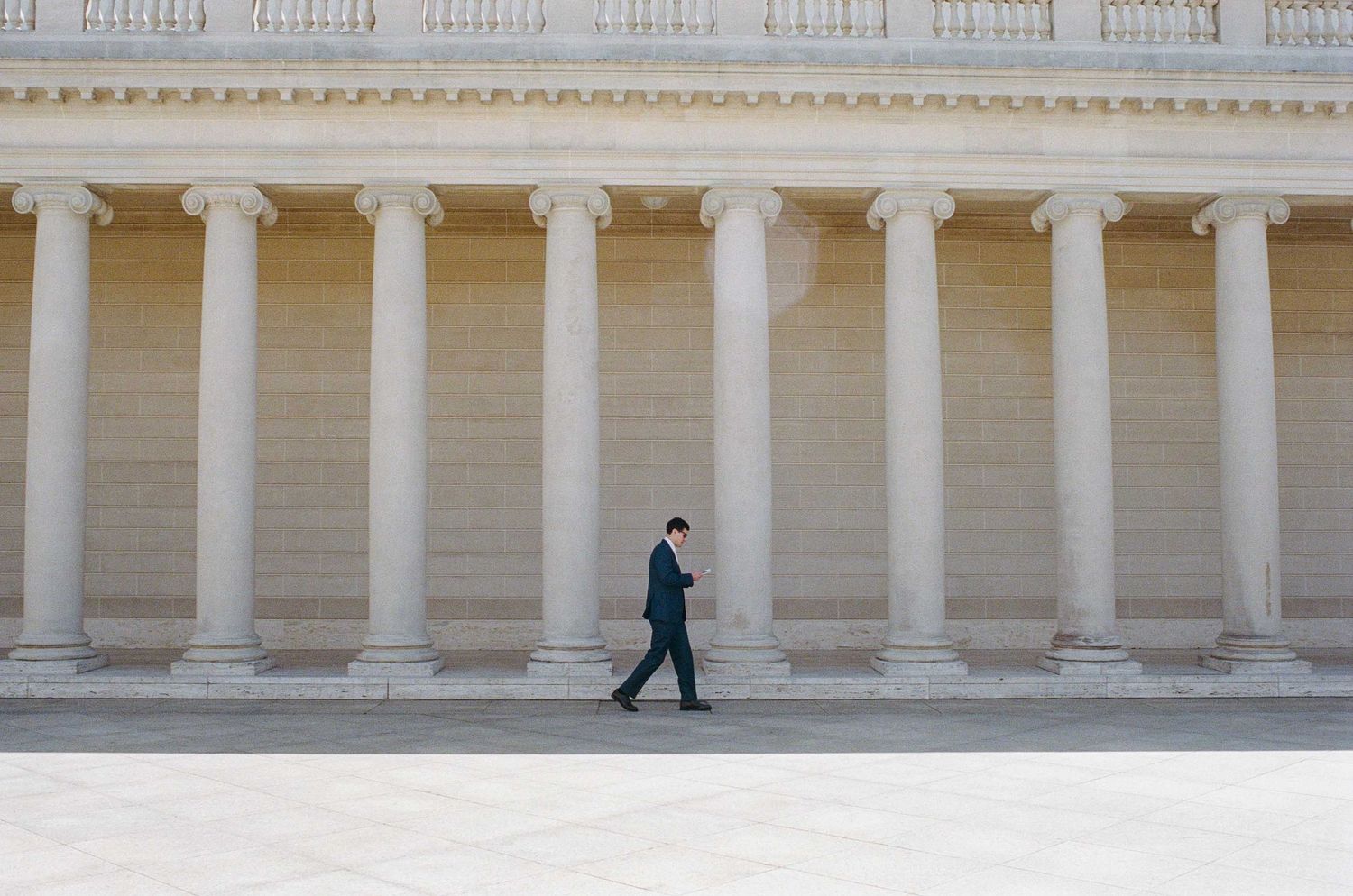 A man in a dark suit walks past grand classical columns of a neoclassical building facade in bright daylight.
