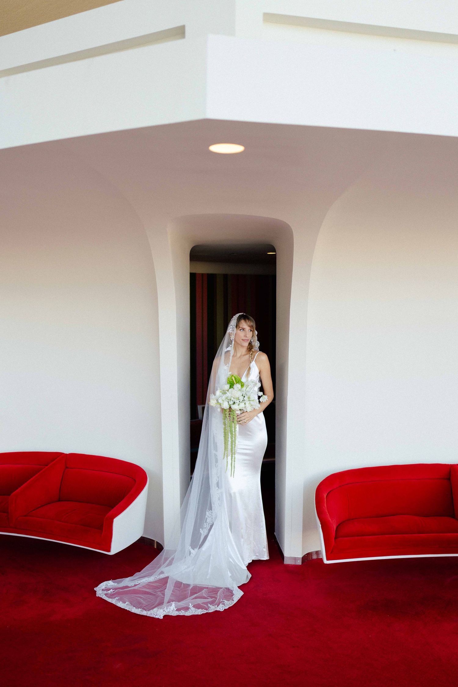 Elegant bride in white satin gown and long veil standing in doorway flanked by two red sofas.