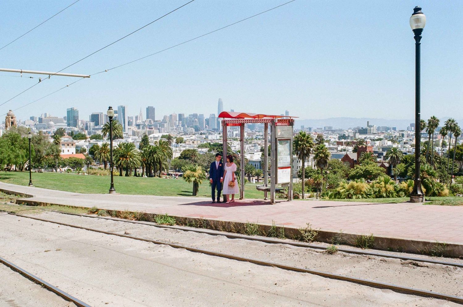 A couple stands at a San Francisco Muni stop with the city skyline visible in the sunny background.