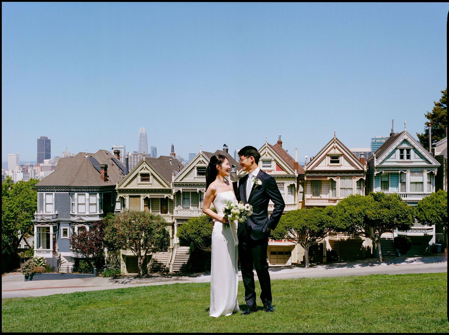 Bride and groom pose at Alamo Square Park with Painted Ladies Victorian houses in background.