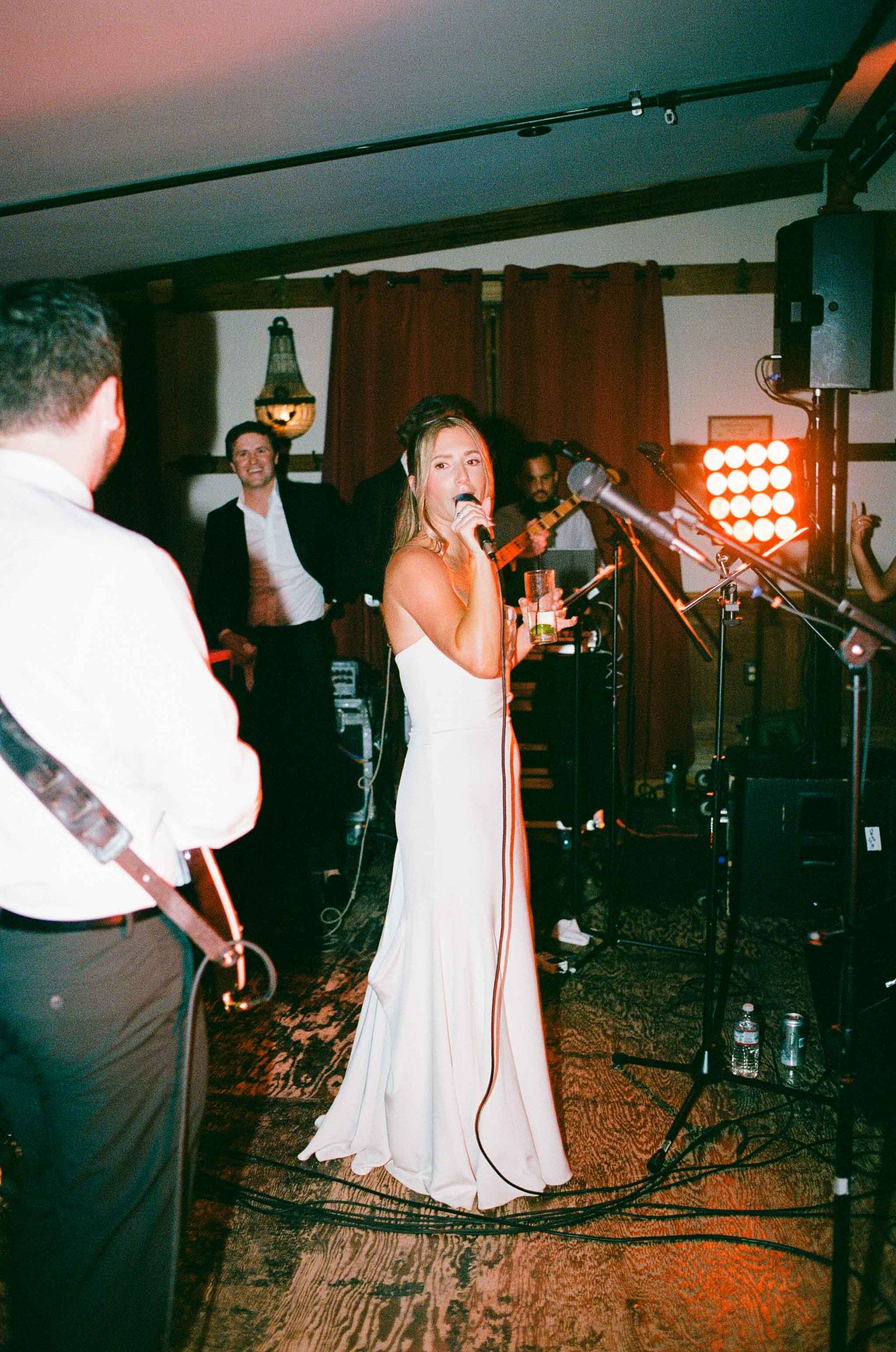 Bride in white gown singing into microphone with live band performing at wedding reception.