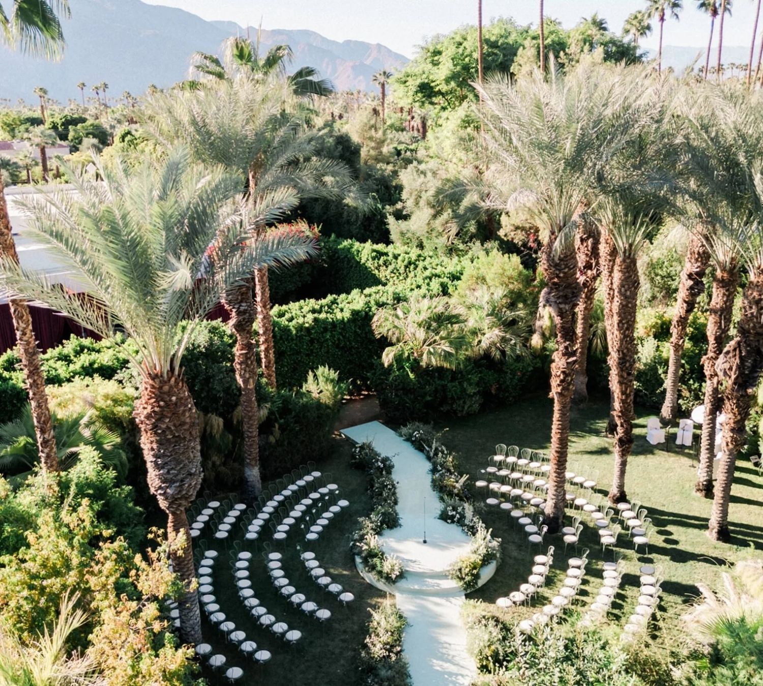 Aerial view of an elegant outdoor wedding ceremony setup with white chairs and palm trees in a lush garden.