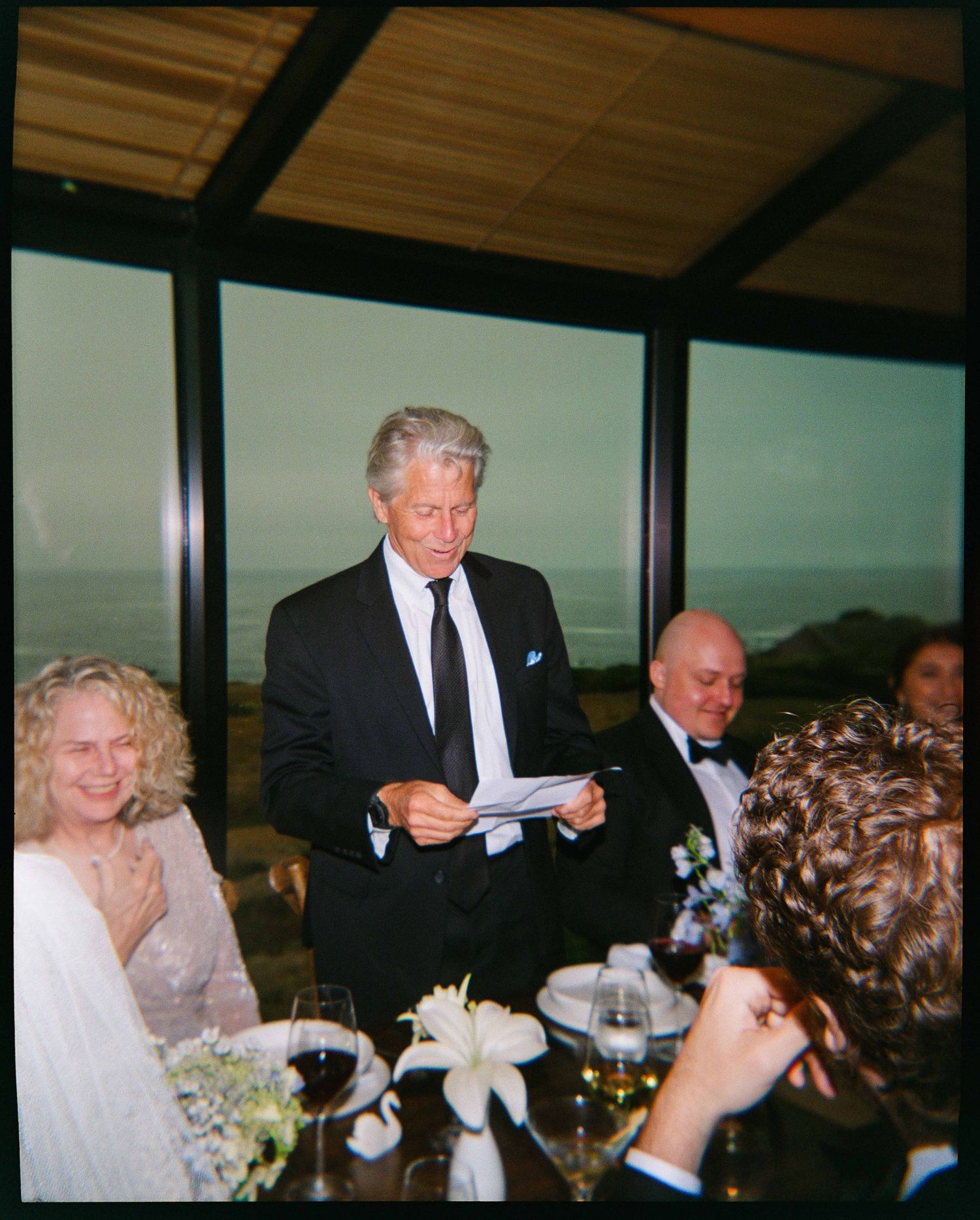 Elegant man in black suit reading notes at formal oceanview dinner party with guests in evening wear.