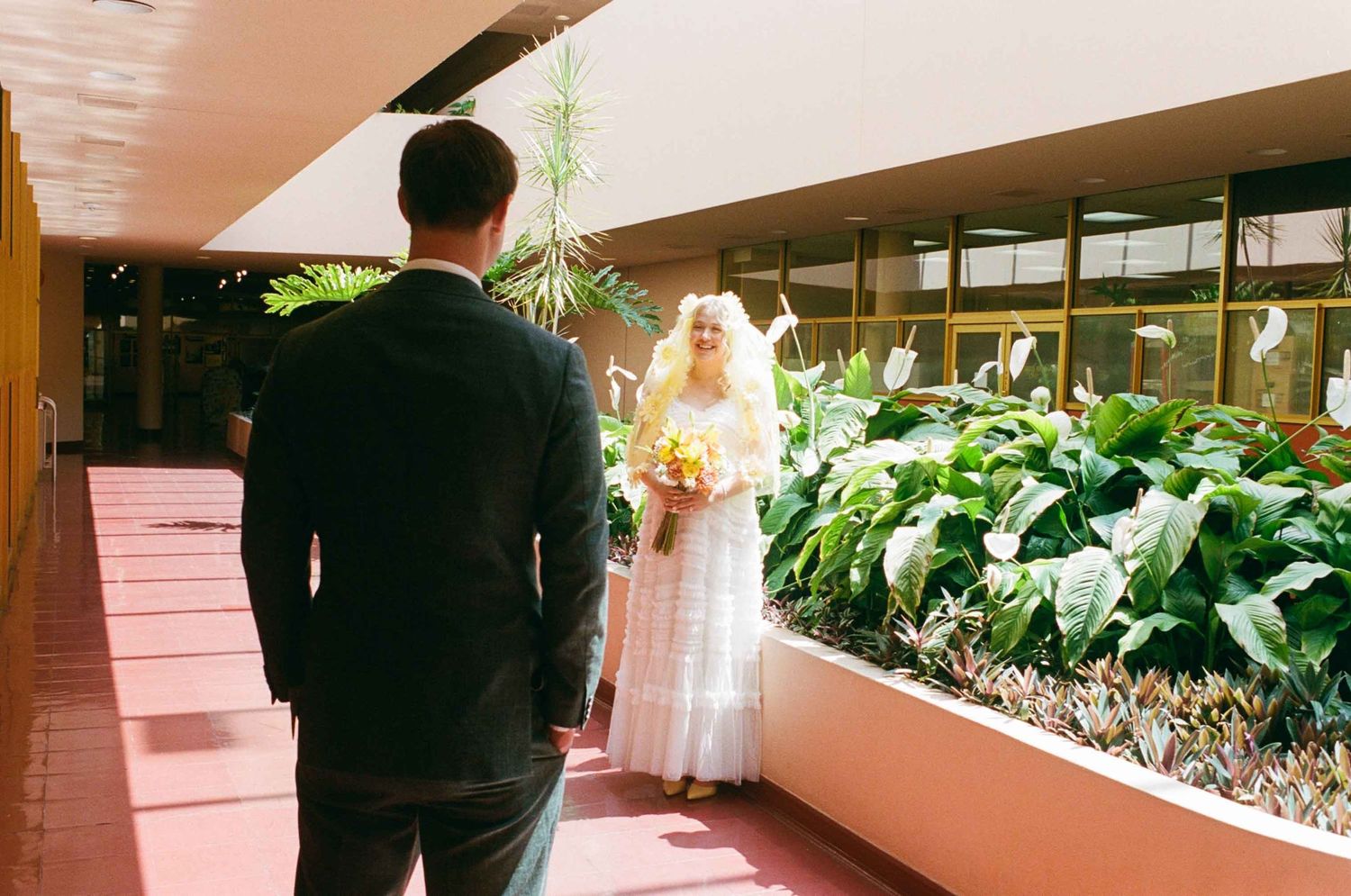 Bride in white dress holding yellow bouquet walks toward groom in black suit along a modern building corridor.