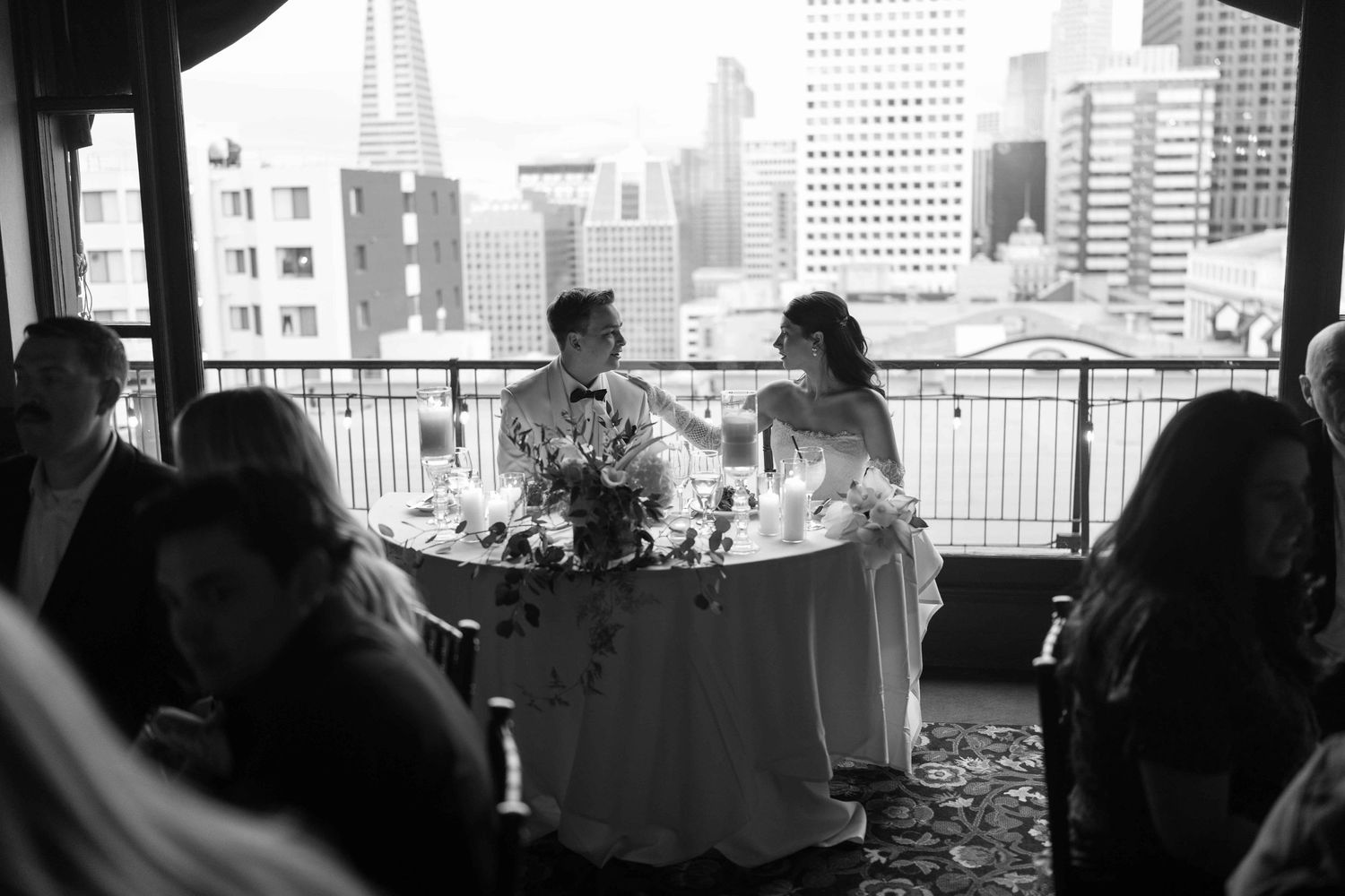 Black and white photo of bride and groom at sweetheart table with city skyline view during reception.