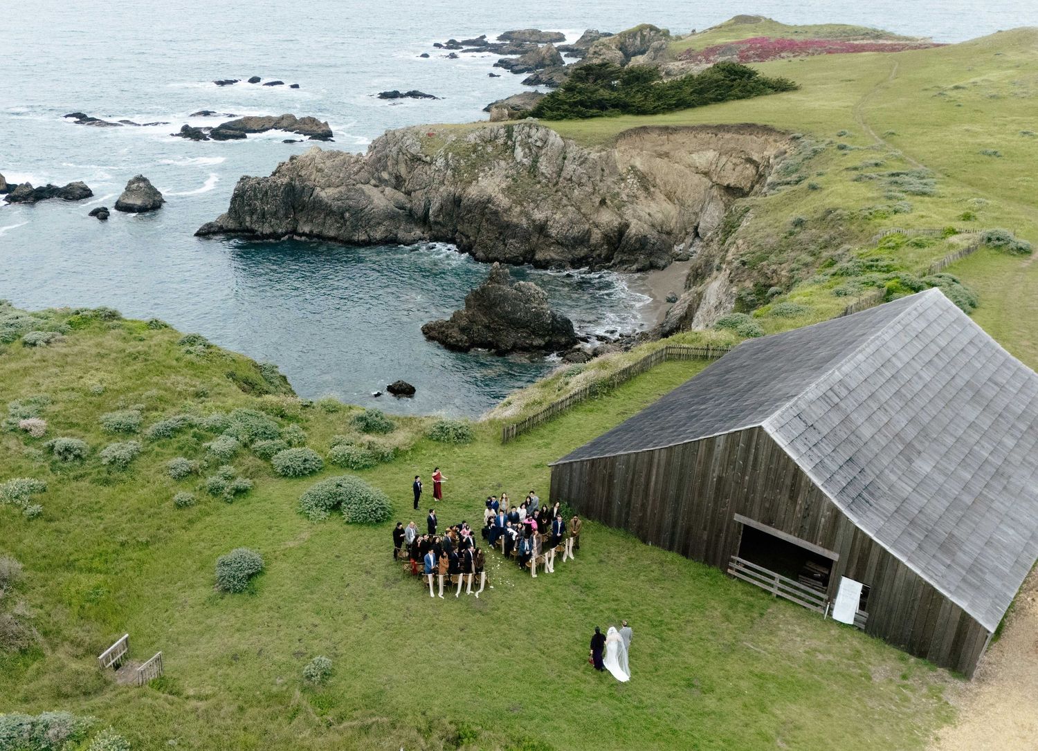 Aerial view of a coastal wedding ceremony near a rustic barn on green cliffs overlooking rocky shores.