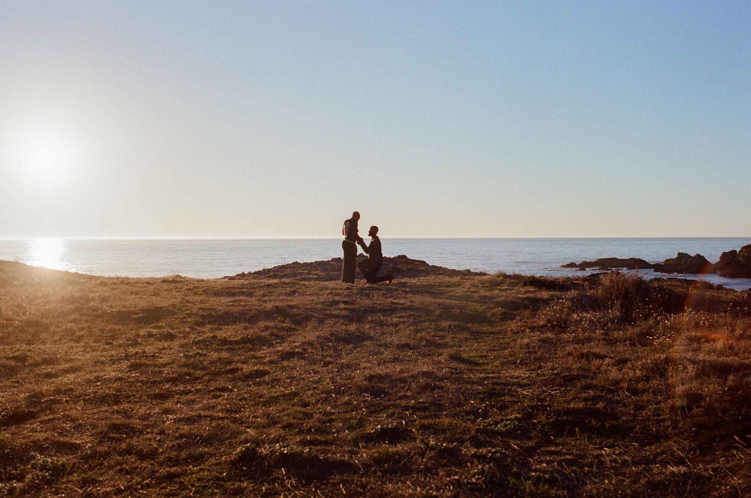 A couple stands together on a coastal cliff at golden hour with the ocean glowing behind them.