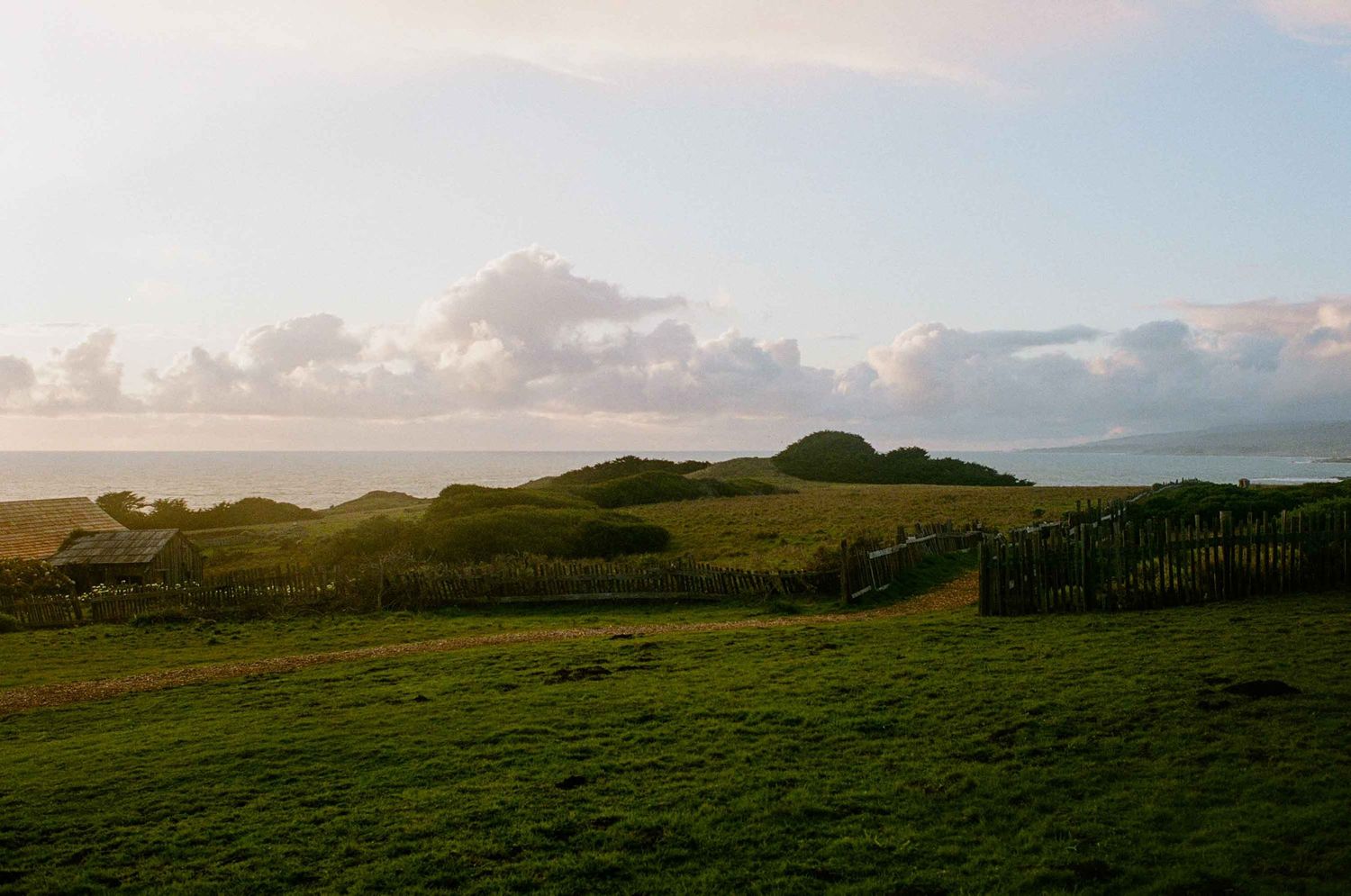 Rolling green hills and grassland beneath a cloudy sunset sky with wooden fencing along the pastoral landscape.
