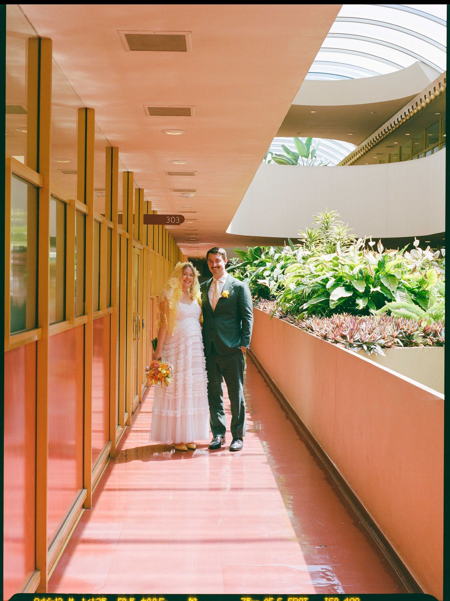Bride and groom walking hand in hand along a modern pink corridor with lush greenery on their wedding day.