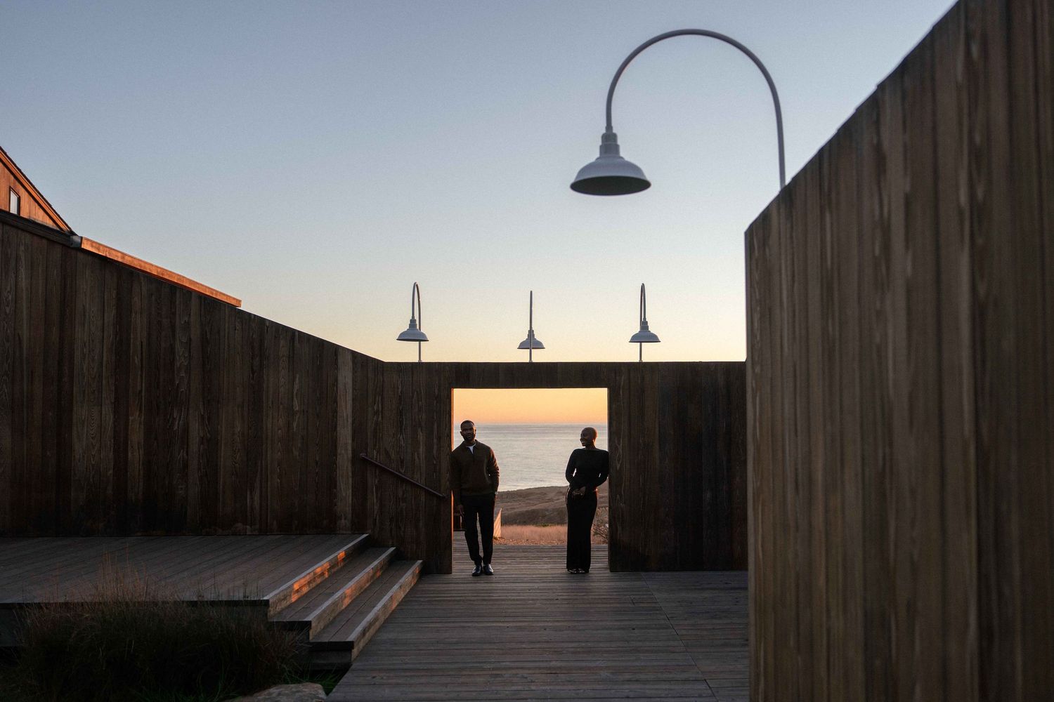 Two people stand on a wooden deck at sunset, framed by tall timber walls and vintage gooseneck lamps overlooking the ocean.