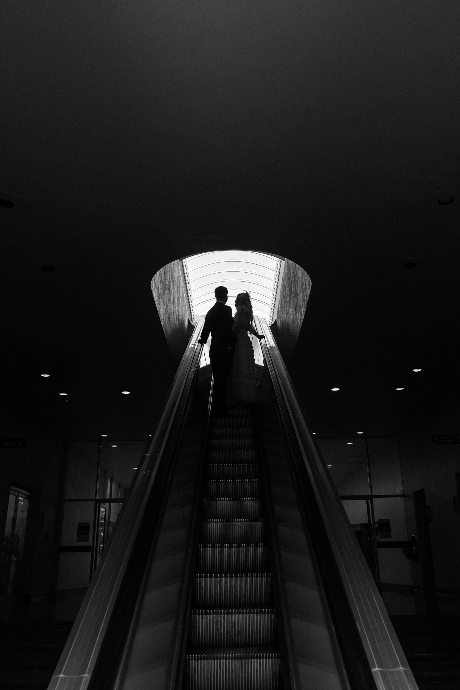 Silhouettes of a couple on an escalator framed by a glowing arch in a dramatic black and white photograph.