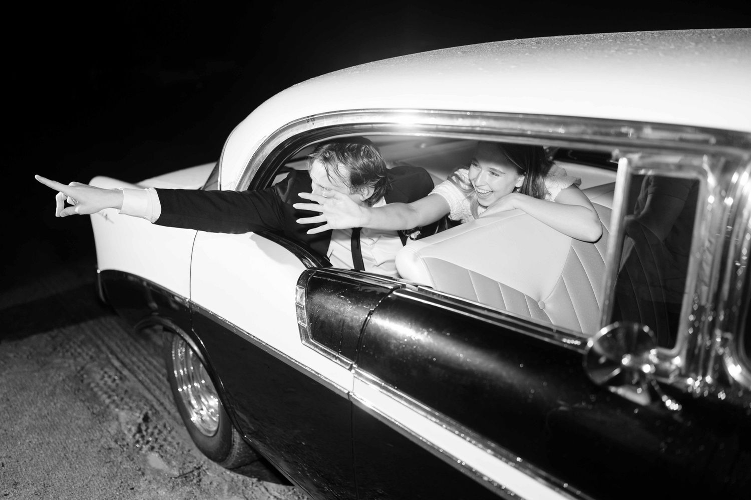 Black and white photo of teens leaning out of a classic 1950s car window at night, pointing into the distance.