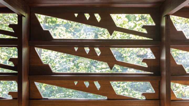 Intricate decorative wooden screen with geometric cutout patterns filtering natural light from surrounding green trees beyond.