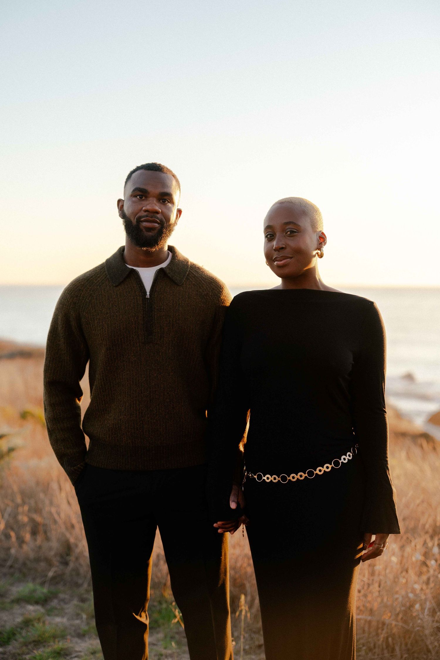 An elegant Black couple poses together at sunset near the ocean in stylish coordinated outfits.