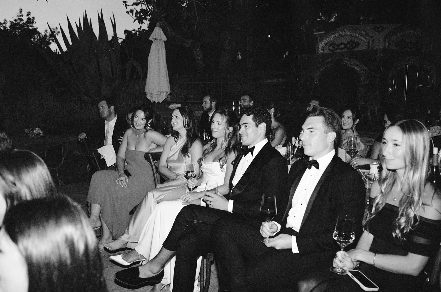 Black and white photo of wedding guests seated outdoors, enjoying drinks at an evening reception.