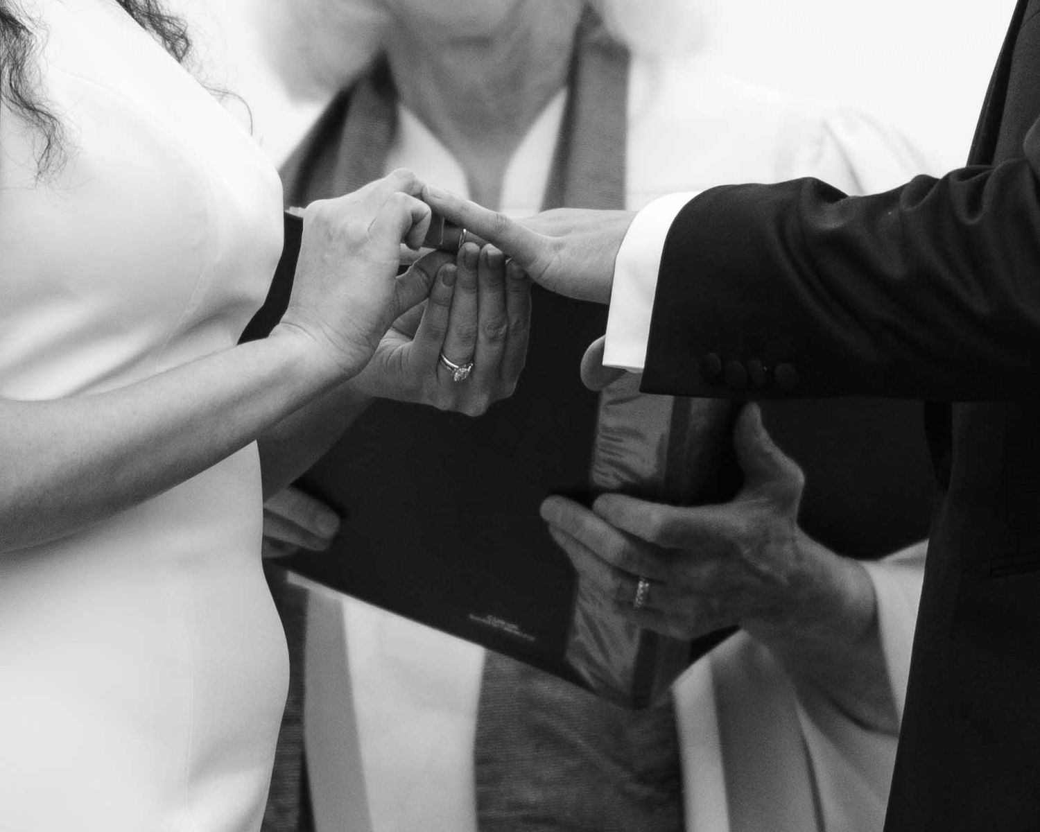 Black and white wedding ceremony photo of couple exchanging rings with officiant holding open book.
