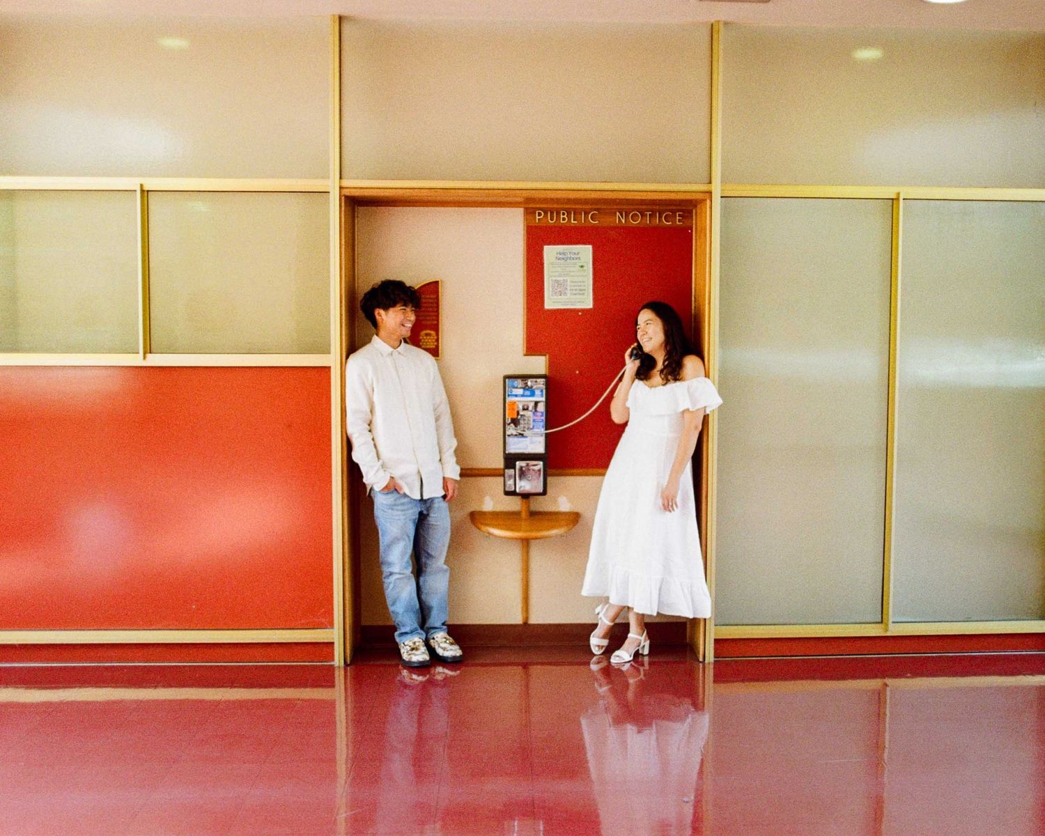 A man in casual wear and a woman in a white dress stand by a payphone in a retro-style lobby.