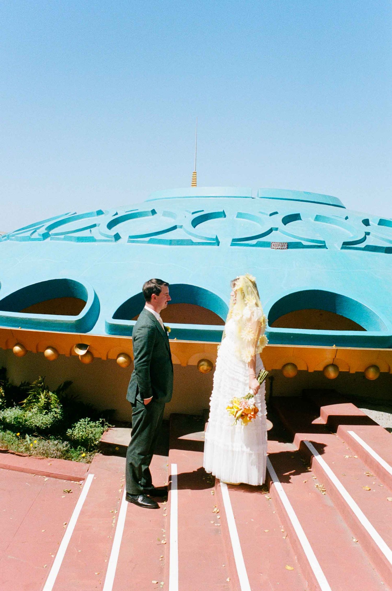 Bride and groom stand before a colorful turquoise retro building on their wedding day.