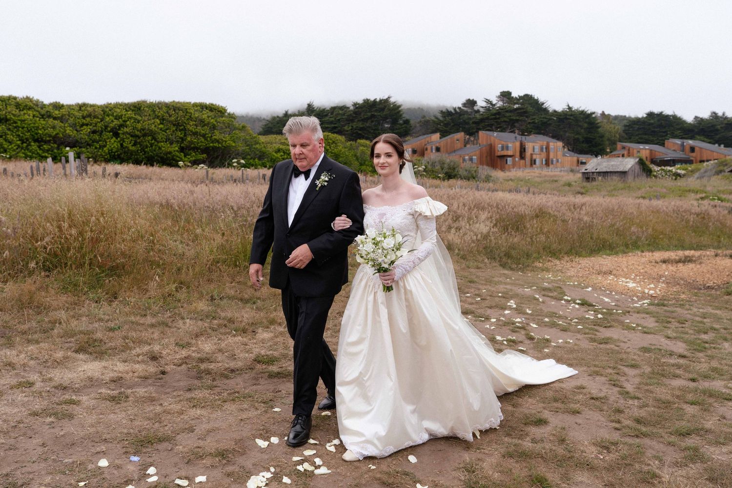Bride in white gown walks with older man in tuxedo on rustic outdoor path with wildflowers and foggy landscape.