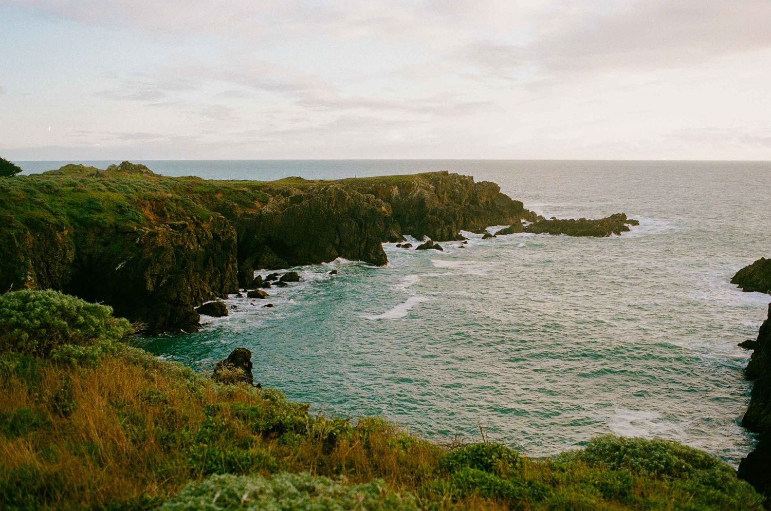 Rocky coastal cliffs covered in lush green vegetation overlook turquoise ocean waters under a cloudy sky.