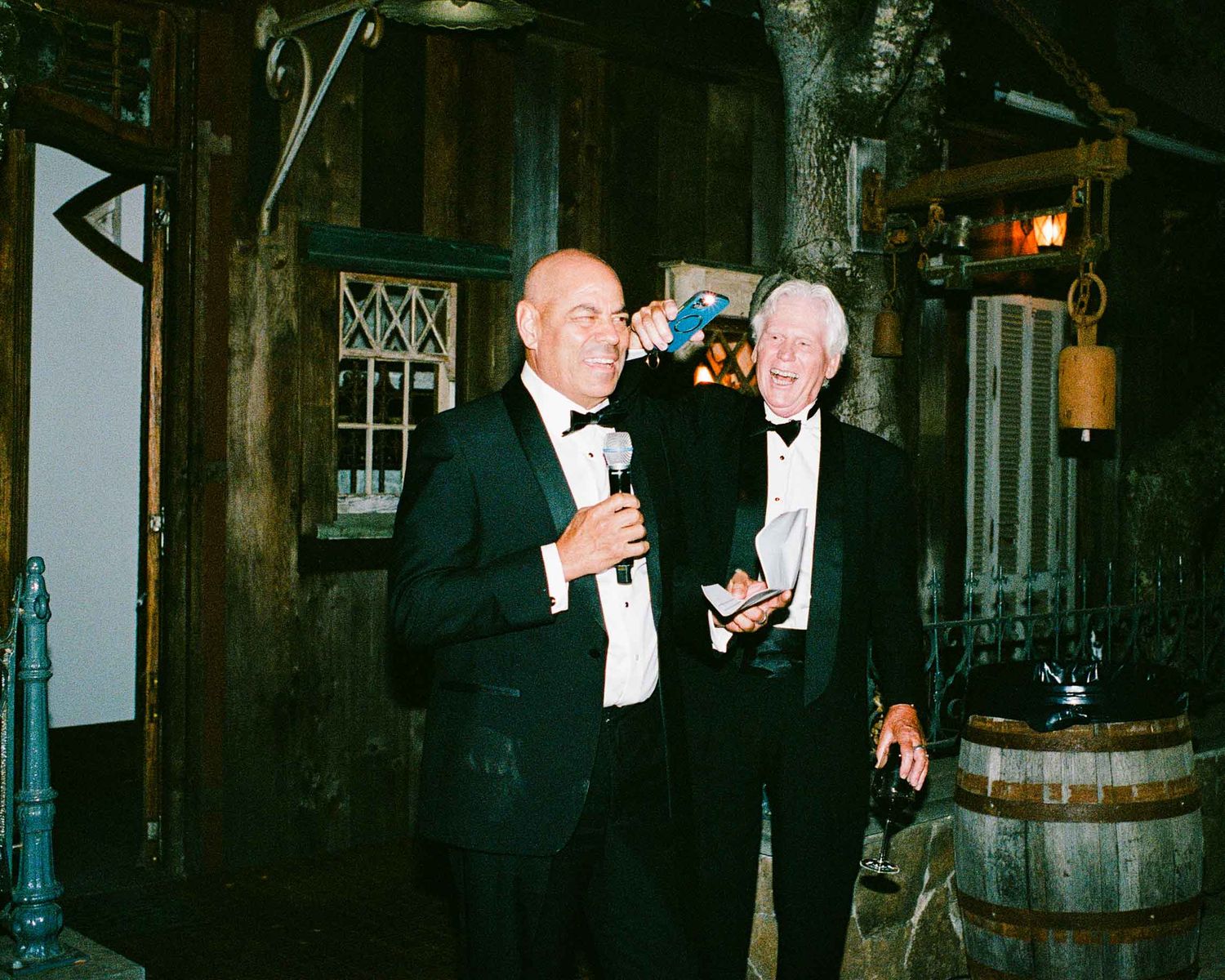 Two men in tuxedos laughing and enjoying drinks inside a rustic wooden wedding venue.