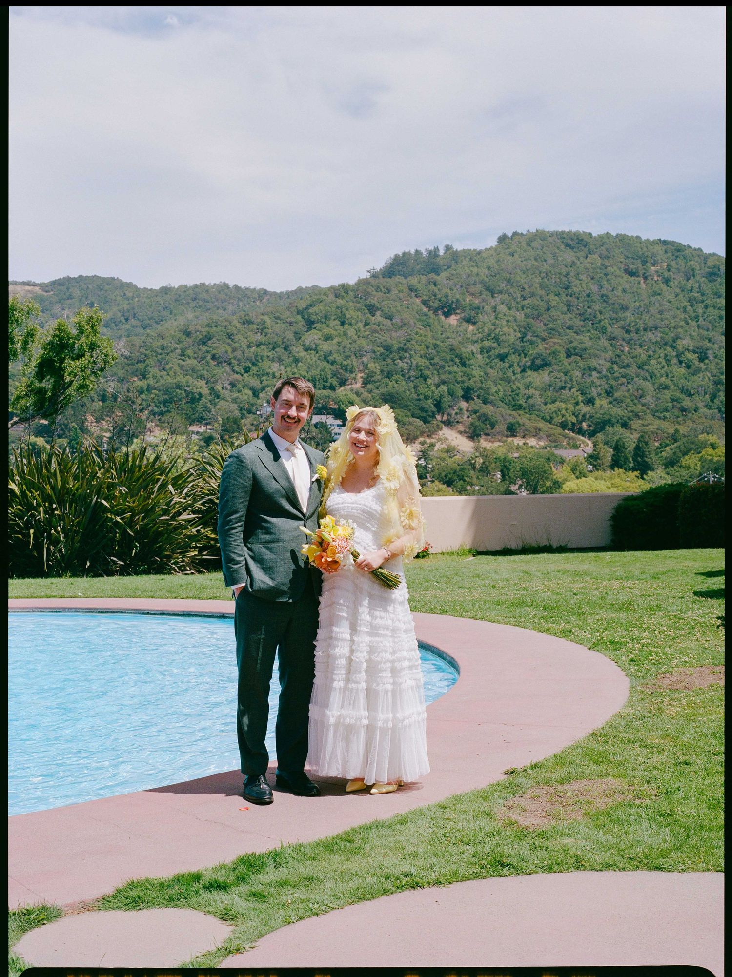 Bride and groom posing by a pool with lush green hills in the background on their wedding day.