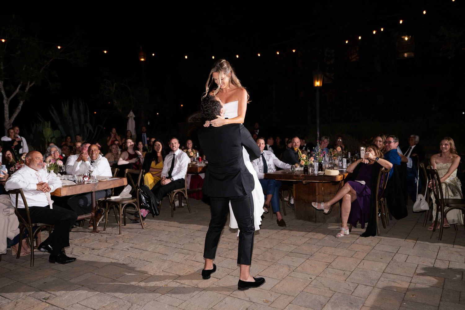 Groom lifting bride joyfully during outdoor evening wedding reception under string lights.