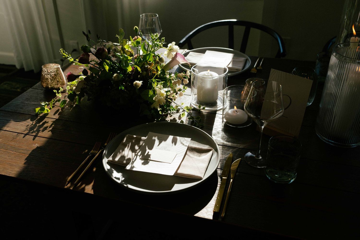 Dramatic moody wedding table setting with floral centerpiece, candle, and elegant place settings in shadow.