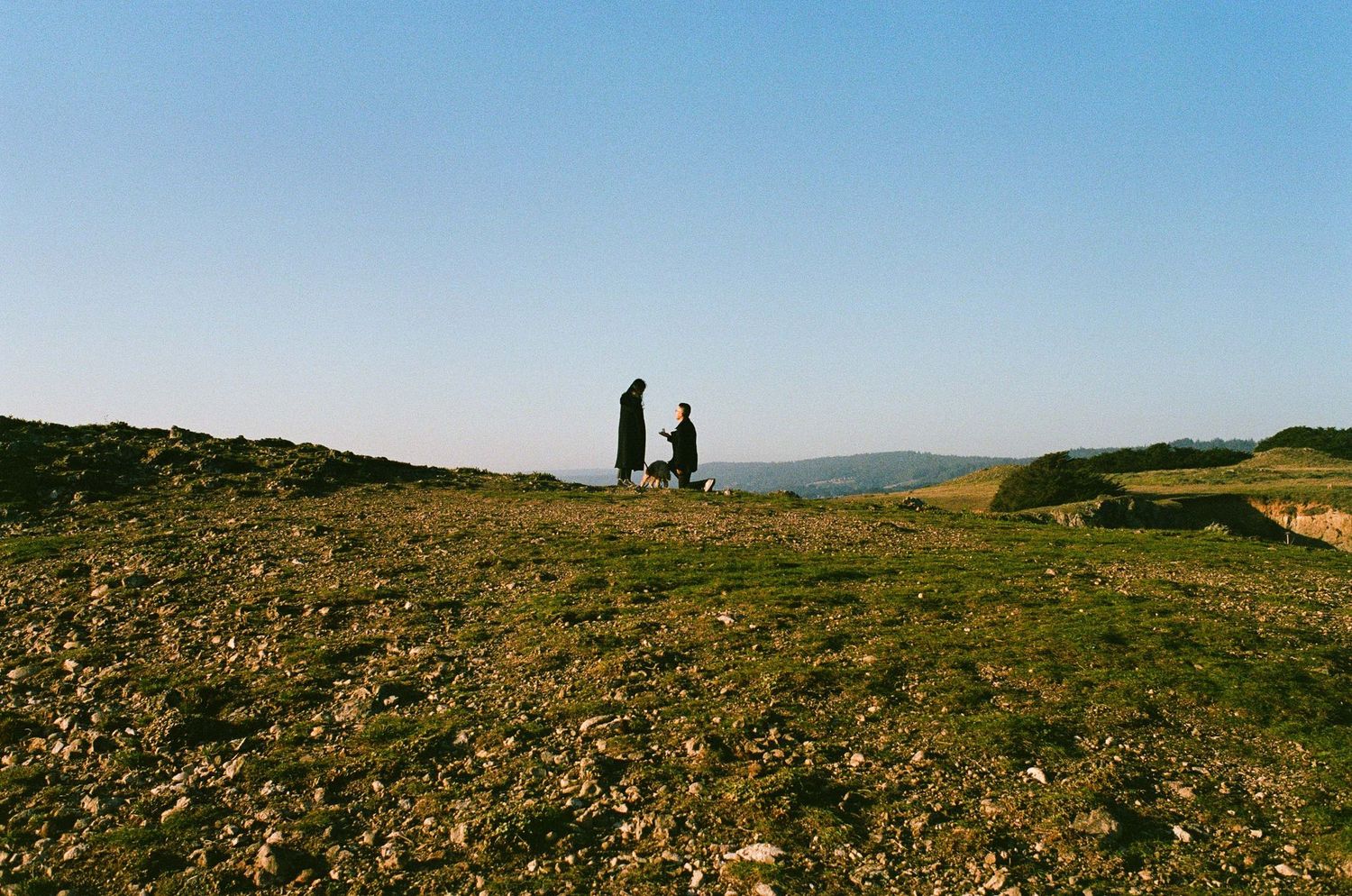 Two people walking a dog on a rocky hilltop landscape under a clear blue sky.