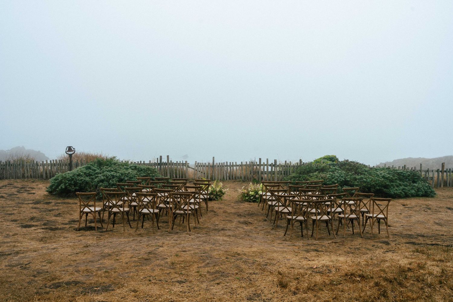 Rustic wooden cross-back chairs arranged in rows for an outdoor wedding ceremony on a foggy coastal field.