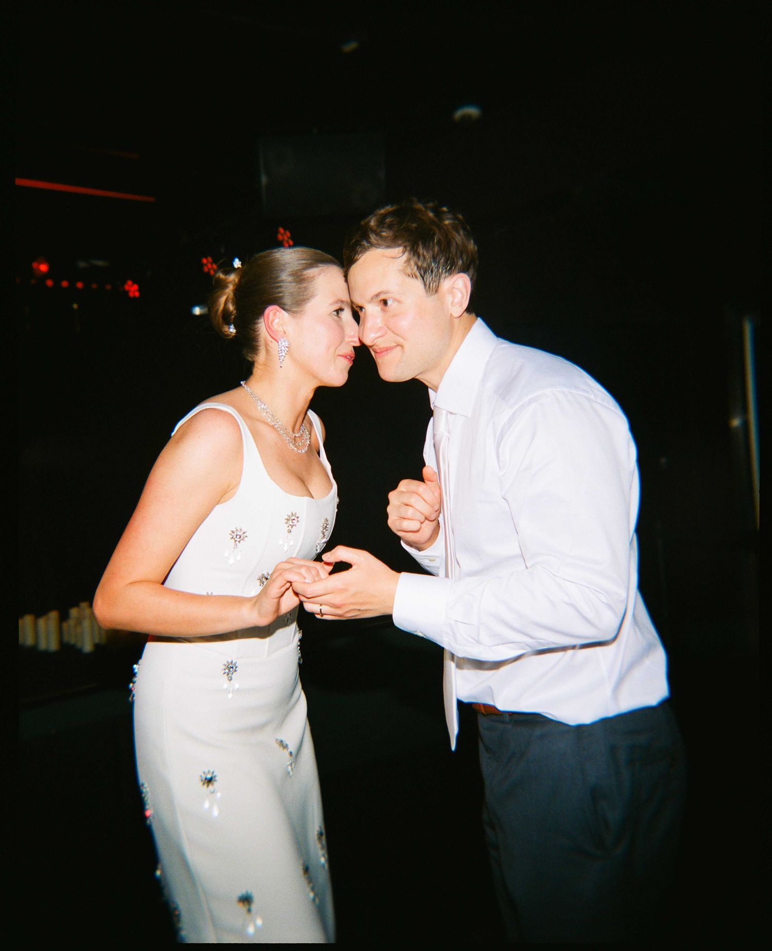 Couple sharing a romantic dance, woman in white floral dress, man in white shirt, at a dimly lit wedding reception.