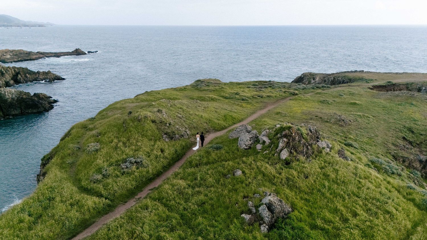Aerial view of a couple walking on a scenic coastal path surrounded by lush green cliffs and ocean views.