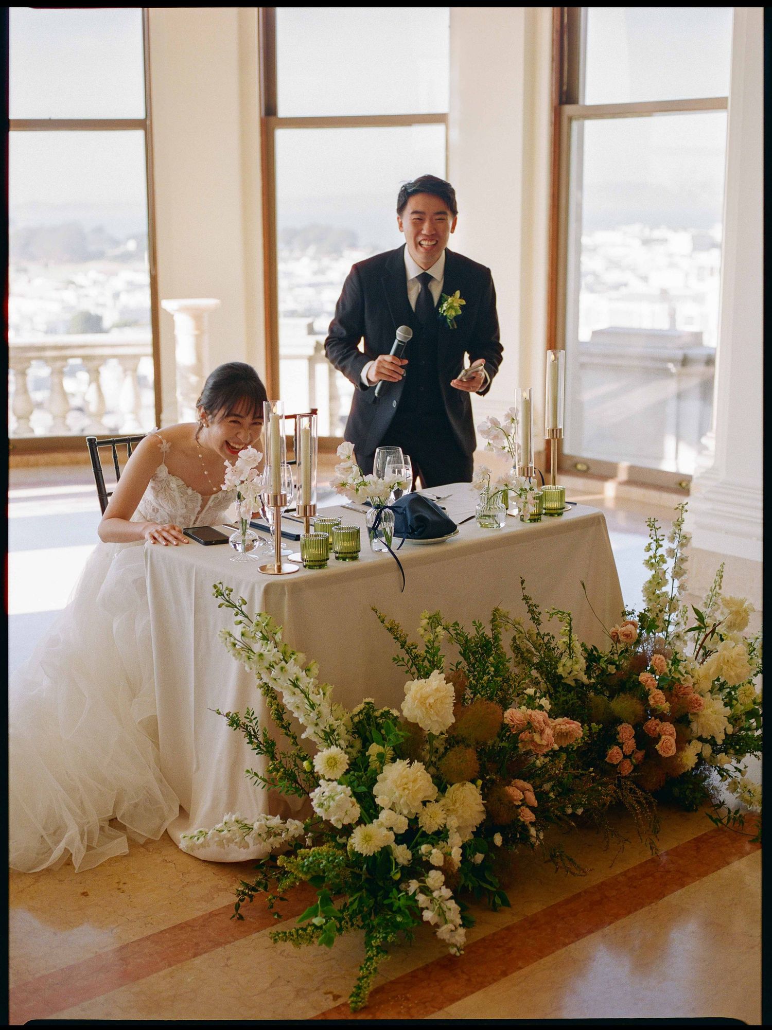 Bride and groom at sweetheart table adorned with white and peach floral arrangements during wedding reception.