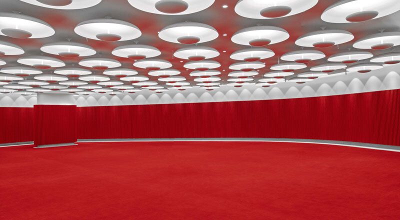 Modern circular hall with red carpet, white geometric ceiling lights, and elegant red curtain walls.