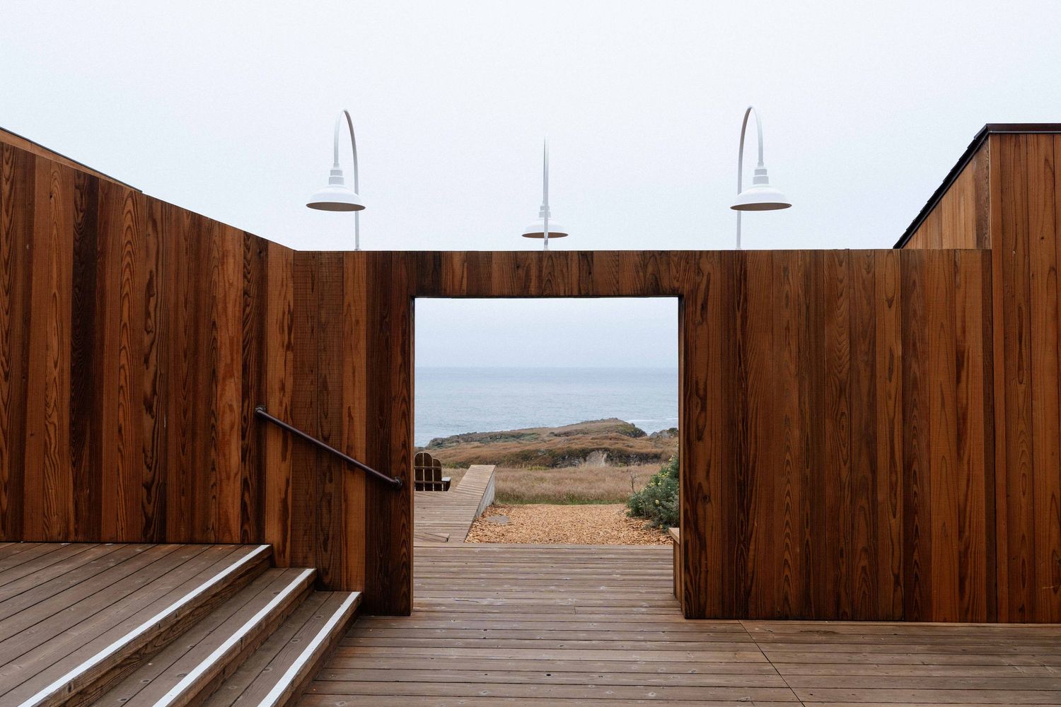 Wooden deck with rich brown timber walls framing an ocean view, featuring two outdoor showers and steps leading to a beach.