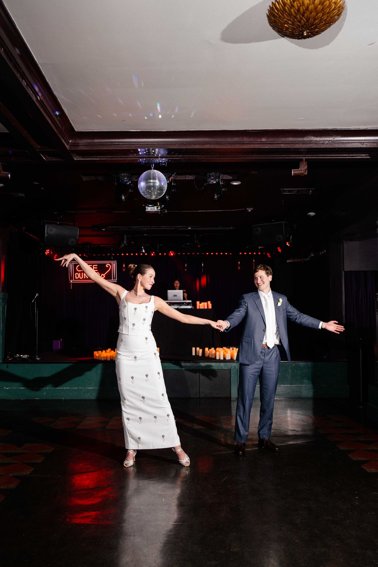 Bride in white gown and groom in blue suit share first dance under disco ball in elegant venue.