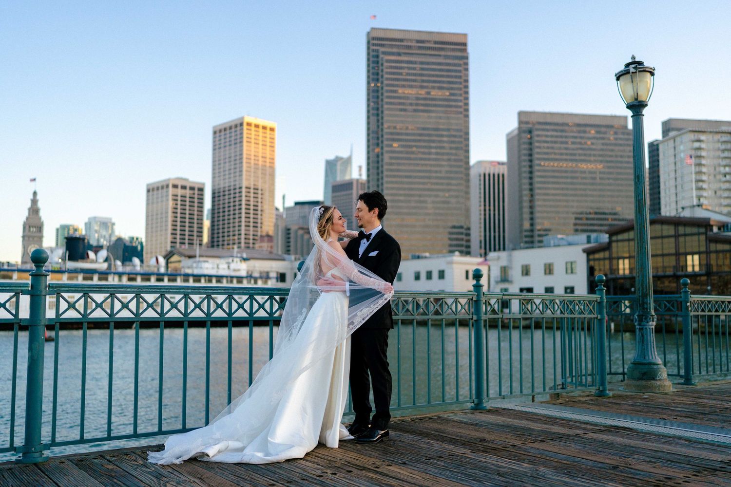A romantic wedding photo against the San Francisco skyline with high-rise buildings and a waterfront railing at dusk.